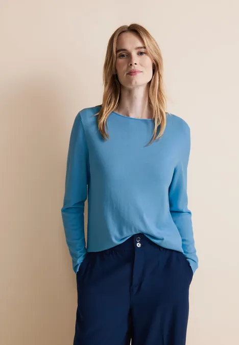 Frau in hellblauem Pullover und dunkler Hose vor beigem Hintergrund.